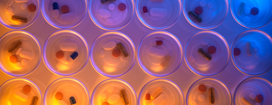 Overhead view of pills and capsules in a grid of clear circular compartments, lit orange on the left and blue on the right.
