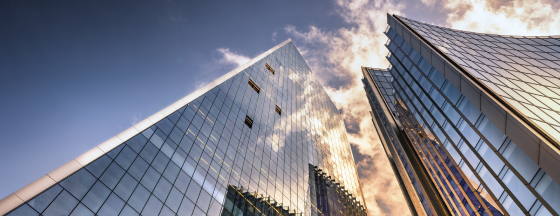 Upward view of modern glass skyscrapers reflecting golden sunlight and clouds against a blue sky.