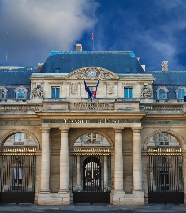 Facade of France's Conseil d'État, featuring stone archways, ornate sculptures, a clock, and French and European Union flags beneath a cloudy blue sky.