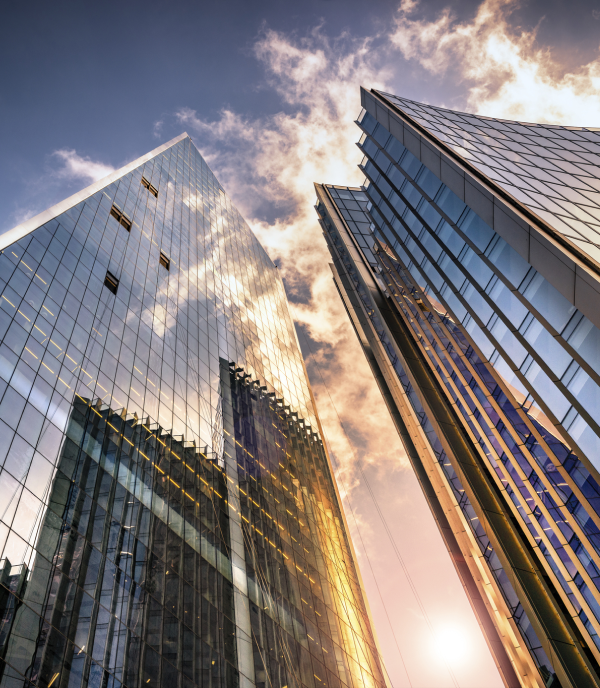 Upward view of modern glass skyscrapers reflecting golden sunlight and clouds against a blue sky.