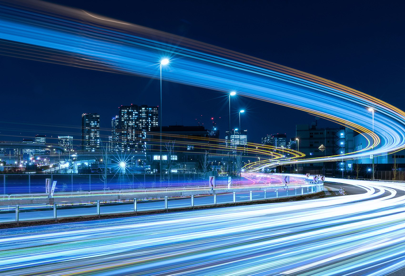 An urban road with high exposure on the vehicles passing. The background has office buildings in the background, the photo was taken at night